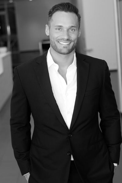 Smiling man in a black suit and white shirt, standing indoors.